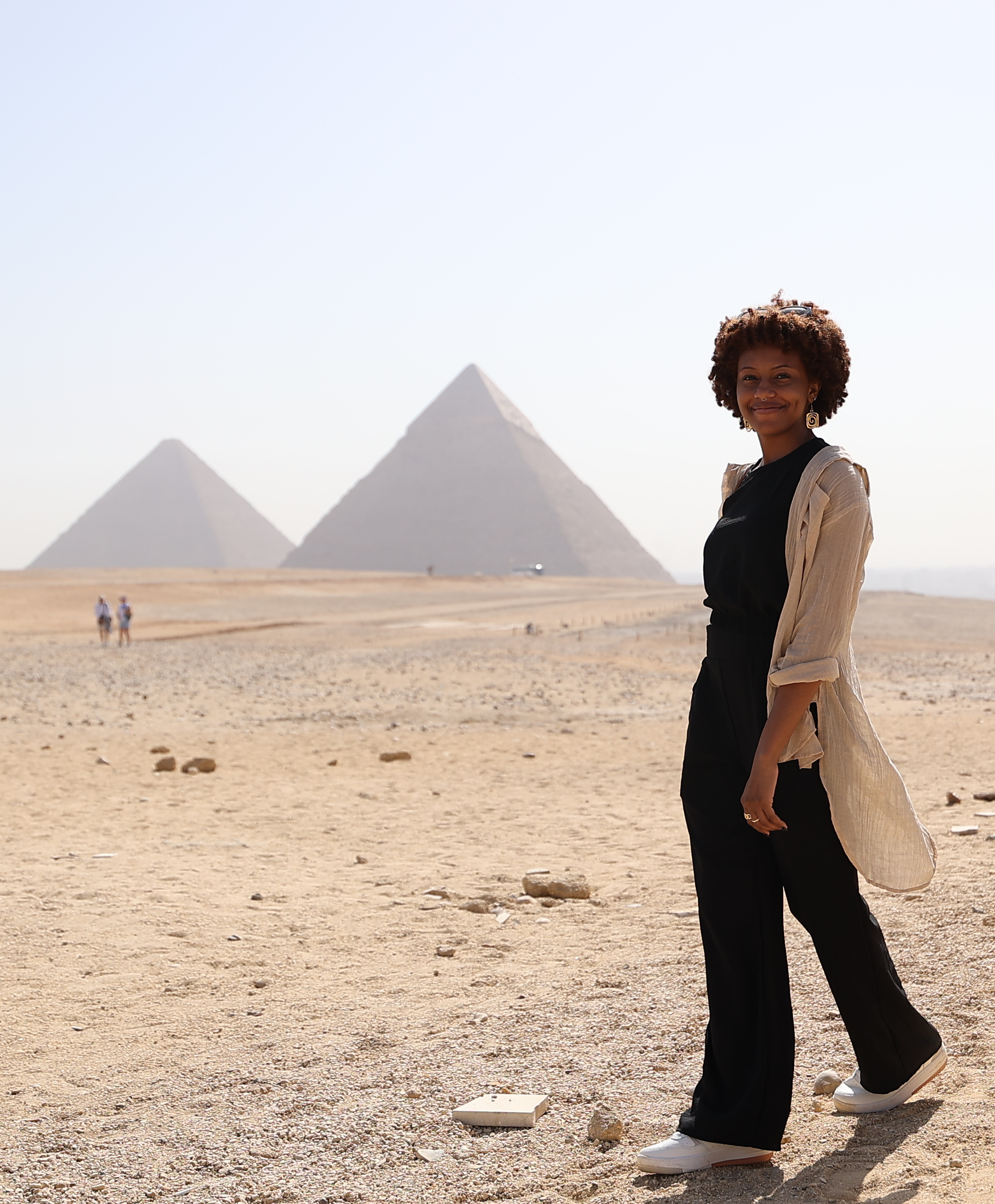 Nia Wright, a black woman with a subtle smile walking in front of 2 pyramids in the desert